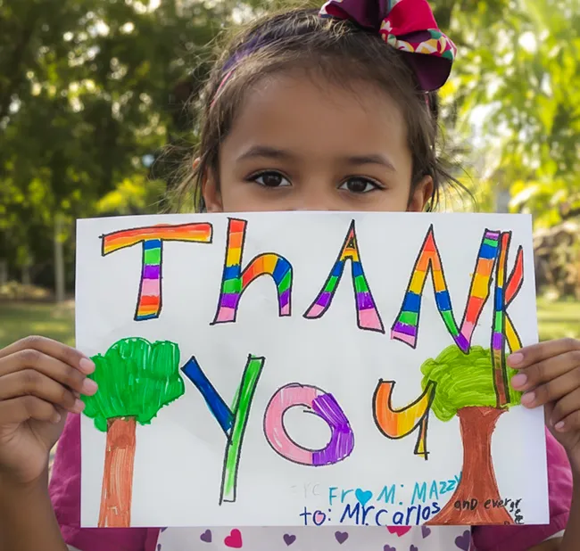 A young girl smiling, holding up a colorful, hand-drawn "THANK YOU" sign from Mazzy to Mr. Carlos and Evergreen, symbolizing community appreciation for tree services.
